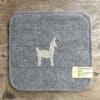Graues Sitzkissen mit Llama-Motiv aus Alpaka- und Schafwolle auf einem Holztisch, Preis und Materialangaben auf einem Etikett.