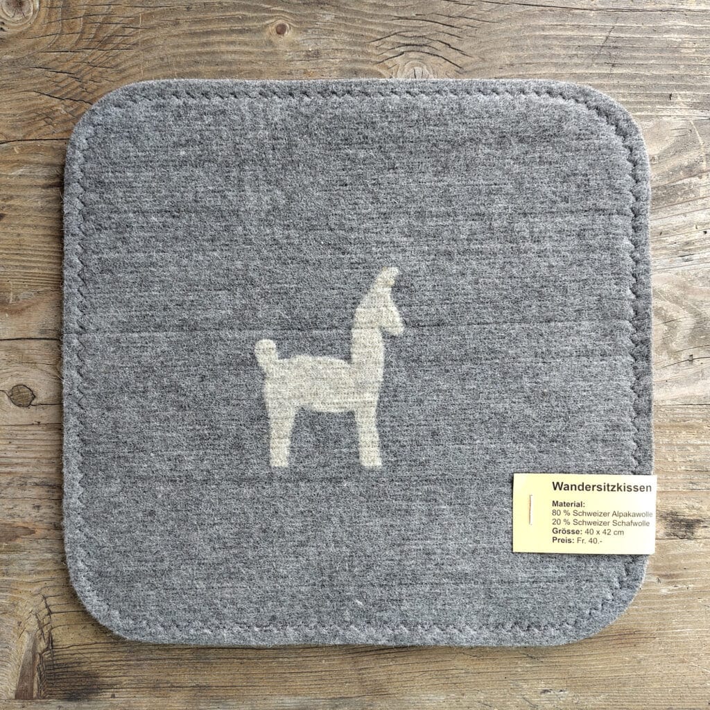 Graues Sitzkissen mit Llama-Motiv aus Alpaka- und Schafwolle auf einem Holztisch, Preis und Materialangaben auf einem Etikett.