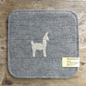 Graues Sitzkissen mit Llama-Motiv aus Alpaka- und Schafwolle auf einem Holztisch, Preis und Materialangaben auf einem Etikett.
