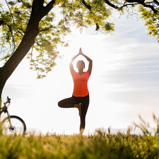 Person in einer Yoga-Pose unter einem Baum im Park, mit einem Fahrrad im Hintergrund.